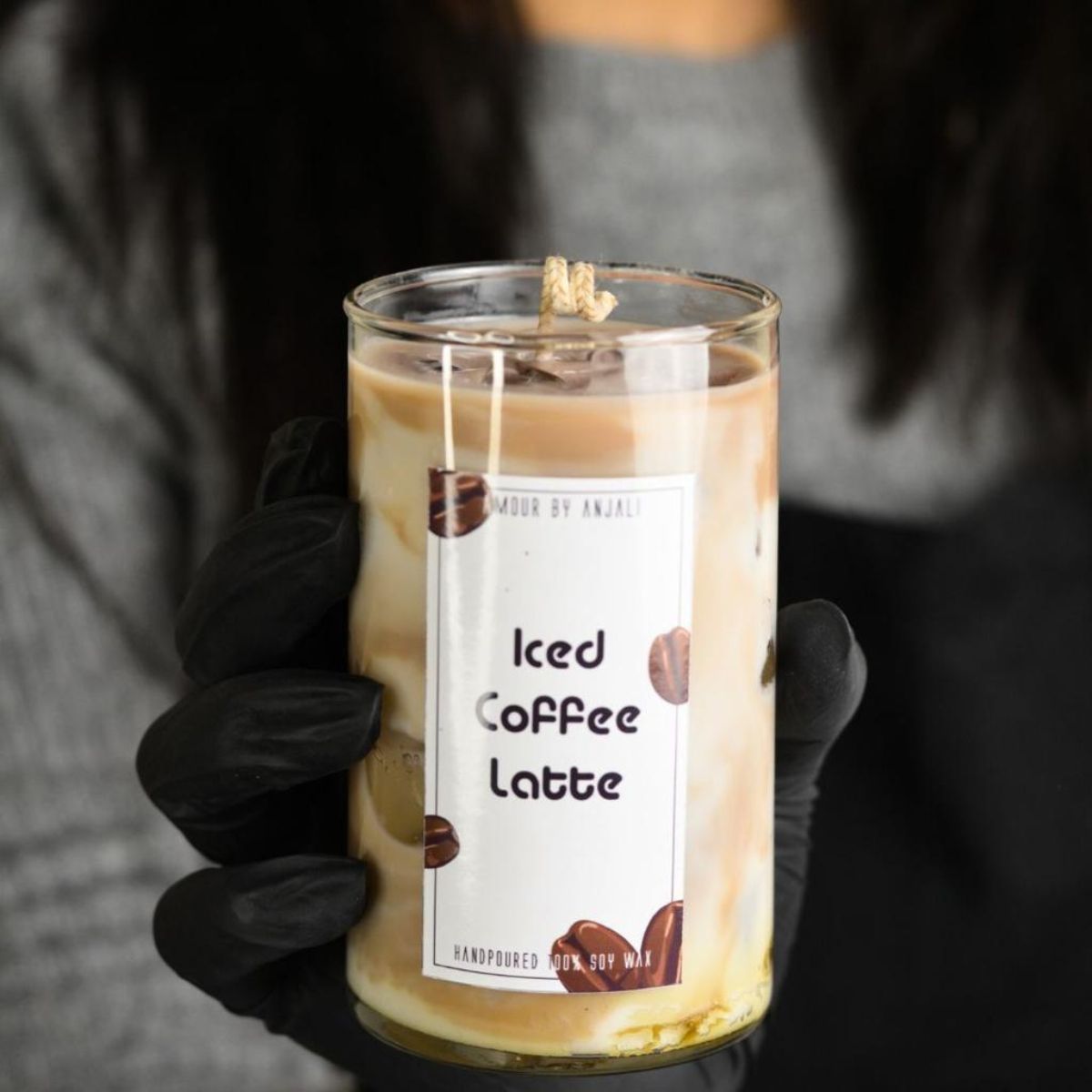 A hand holding a candle with a label that reads 'Iced Coffee Latte', set against a blurred background.