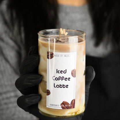 A hand holding a candle with a label that reads 'Iced Coffee Latte', set against a blurred background.