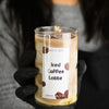 A hand holding a candle with a label that reads 'Iced Coffee Latte', set against a blurred background.